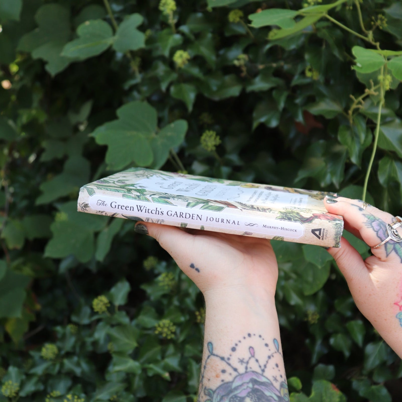 Someone holding the Green Witch's Garden Journal by Arin Murphy-Hiscock in front of greenery, showing off the side.