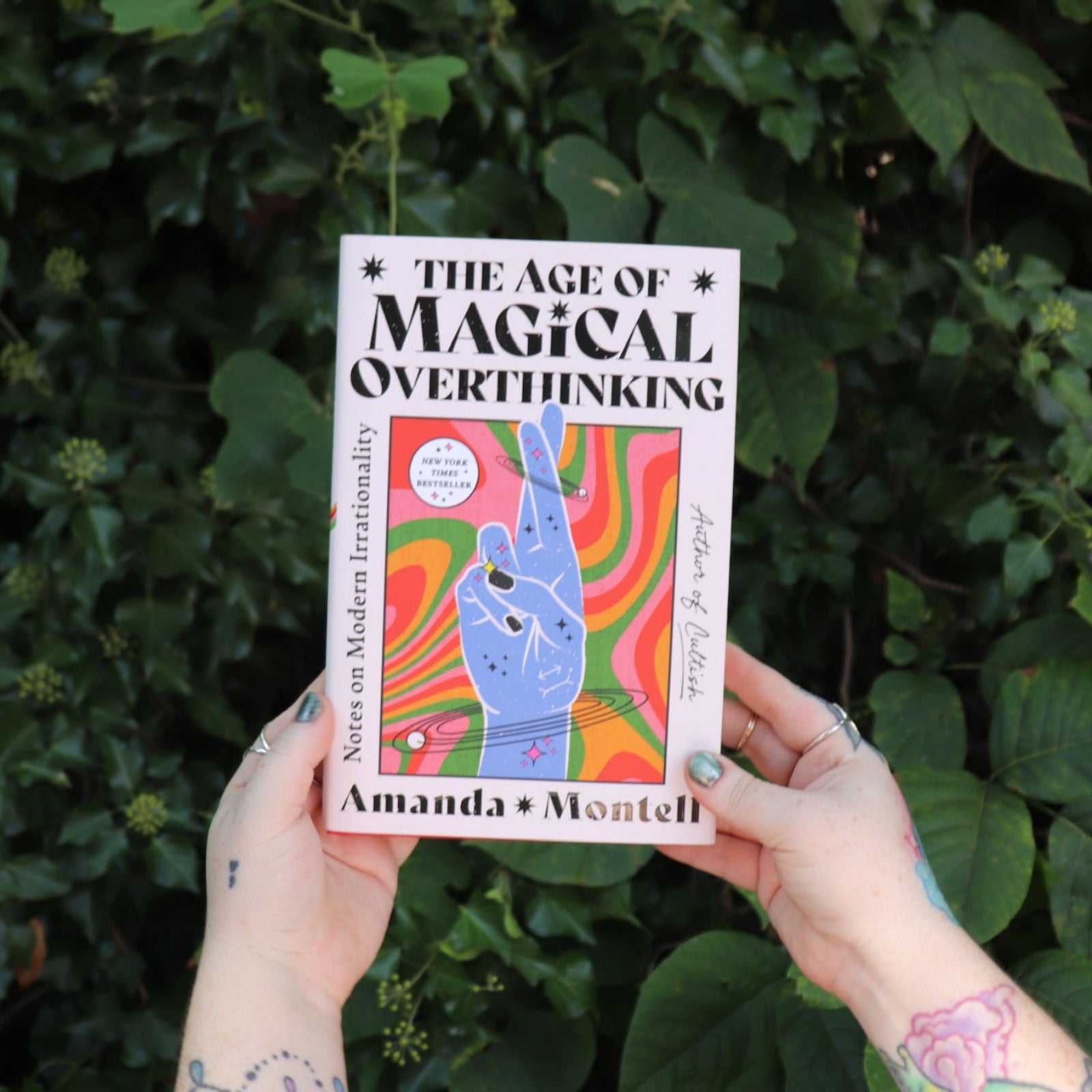 Someone holding the Age of Magical Overthinking Hardcover Book in front of greenery.