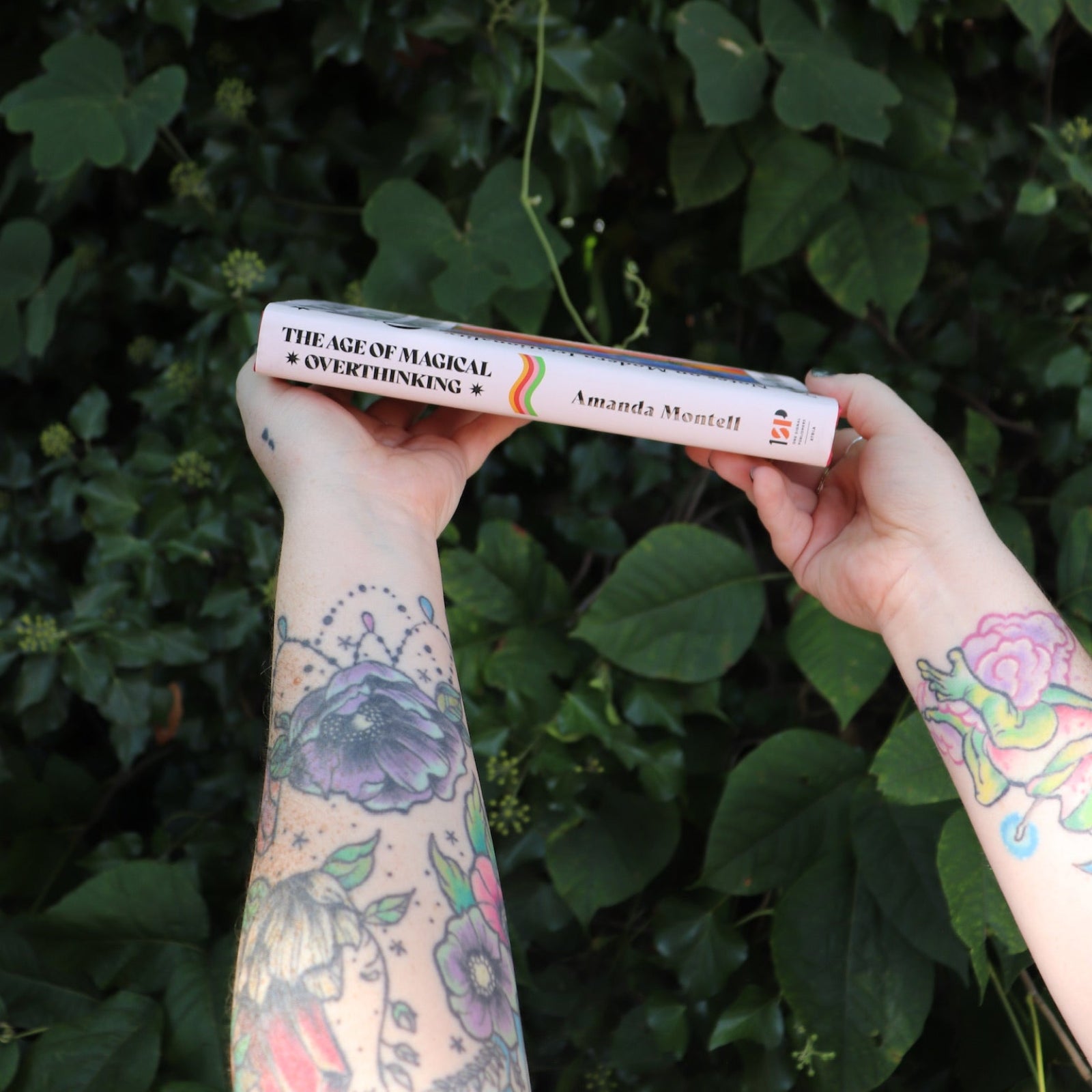 Someone holding the Age of Magical Overthinking Hardcover Book in front of greenery, showing off the side.