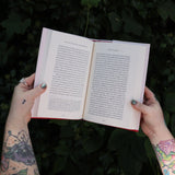 Someone holding the Age of Magical Overthinking Hardcover Book open in front of greenery, showing off the pages.