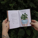 Someone holding the Houseplants and Their F* Up Thoughts by Carlyle Christoff open in front of greenery, showing off the pages.