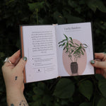 Someone holding the Houseplants and Their F* Up Thoughts by Carlyle Christoff open in front of greenery, showing off the pages.