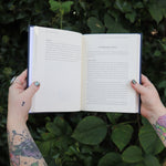 Someone holding the Spells for Change Hardcover Book open in front of greenery, showing off the pages.