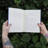 Someone holding the Spells for Change Hardcover Book open in front of greenery, showing off the pages.