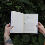 Someone holding the Spells for Change Hardcover Book open in front of greenery, showing off the pages.