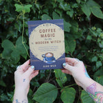 Someone holding the Coffee Magic for the Modern Witch Paperback Book in front of greenery.