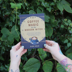 Someone holding the Coffee Magic for the Modern Witch Paperback Book in front of greenery.