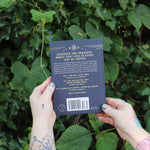 Someone holding the Coffee Magic for the Modern Witch Paperback Book in front of greenery, showing off the side.