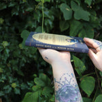 Someone holding the Coffee Magic for the Modern Witch Paperback Book in front of greenery, showing off the side.