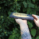 Someone holding the Coffee Magic for the Modern Witch Paperback Book in front of greenery, showing off the side.