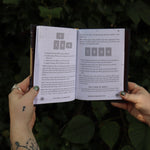 Someone holding the Coffee Magic for the Modern Witch Paperback Book open in front of greenery, showing off the pages.