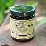 The back of the 7 Medicinal Mushrooms + Cacao Adaptogenic 4 oz Powder on a rusty red surface surrounded by green ivy.