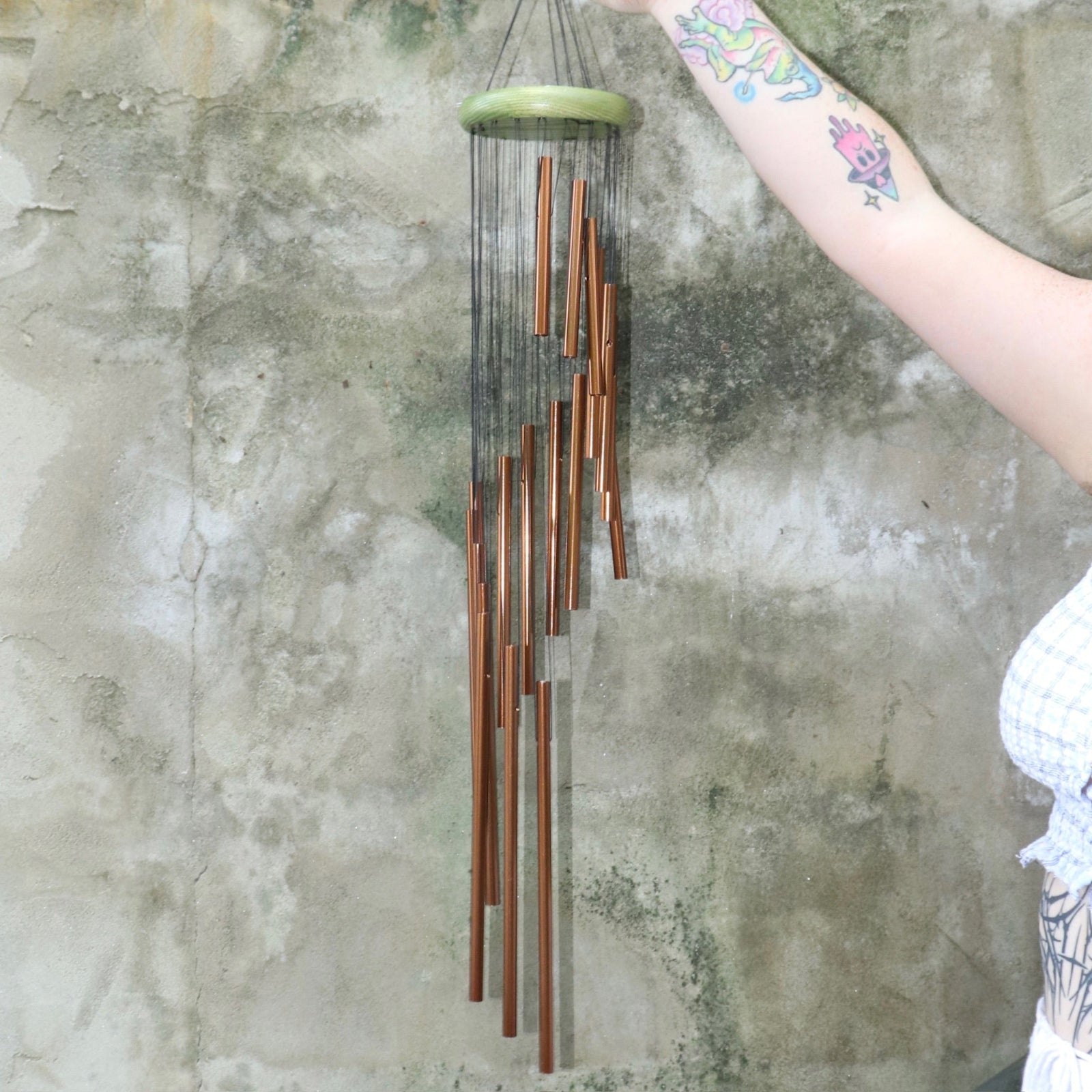 Someone holding the Green Woodstock Habitats™ Rainfall Wind Chime up in front of a concrete wall.