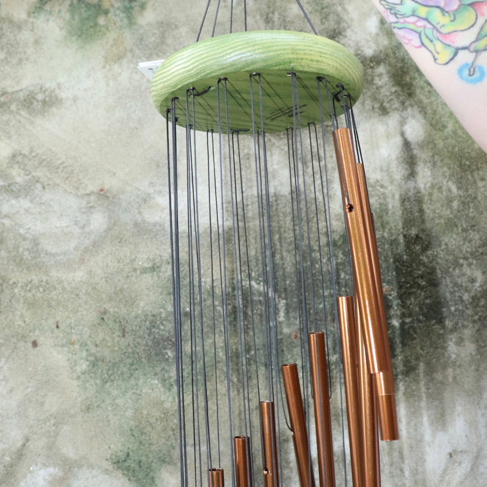 An up close image of someone holding the Green Woodstock Habitats™ Rainfall Wind Chime up in front of a concrete wall.