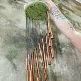 An up close image of someone holding the Green Woodstock Habitats™ Rainfall Wind Chime up in front of a concrete wall.