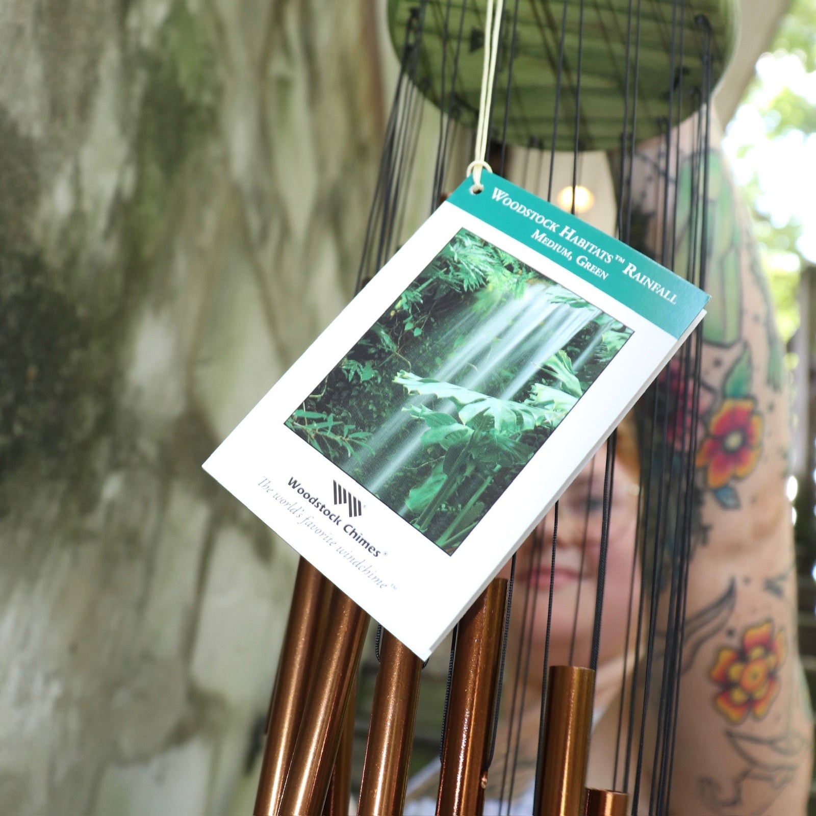 An up close image of someone holding the Green Woodstock Habitats™ Rainfall Wind Chime up in front of a concrete wall.