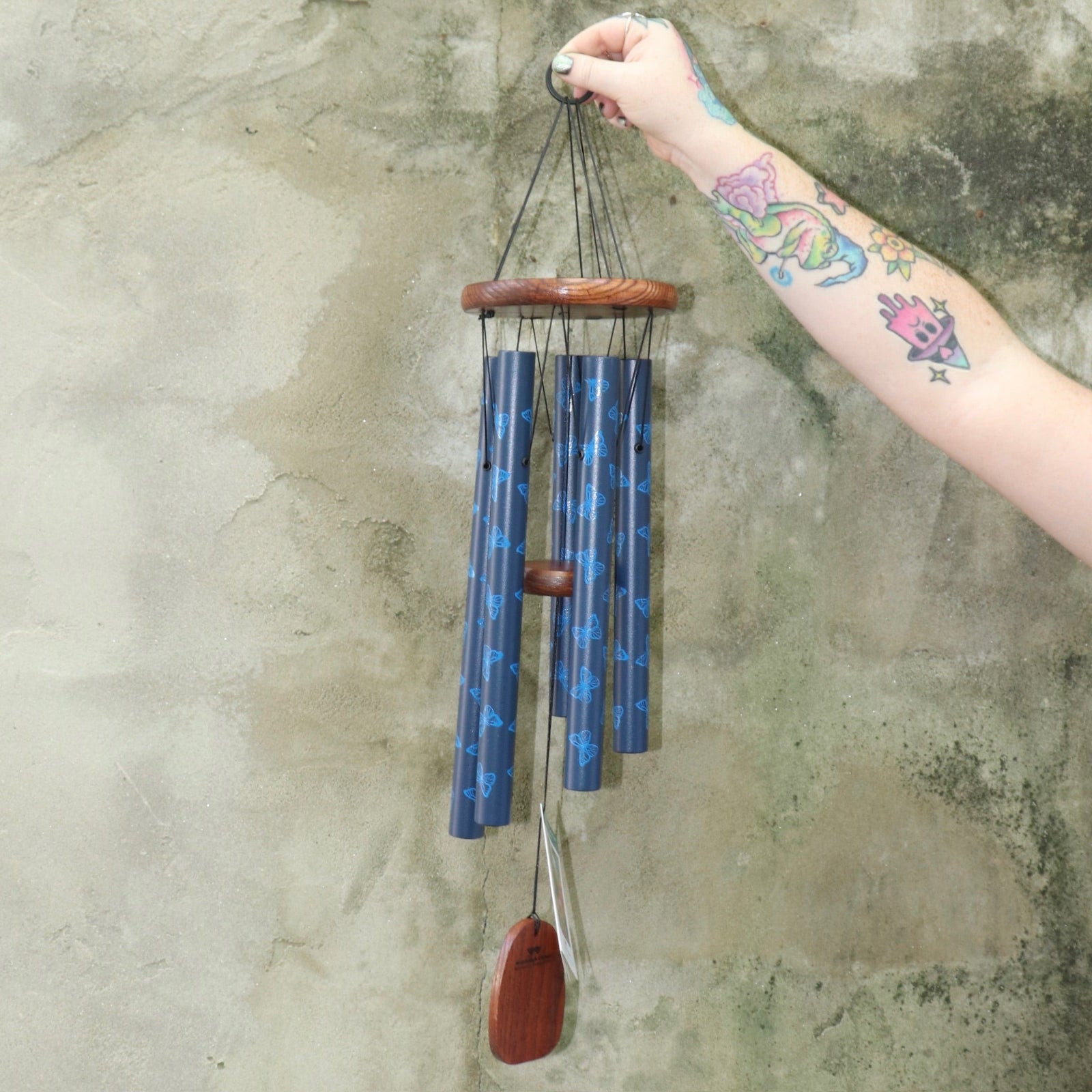Someone holding the Butterfly Woodstock Garden Chime™ in front of a concrete wall.