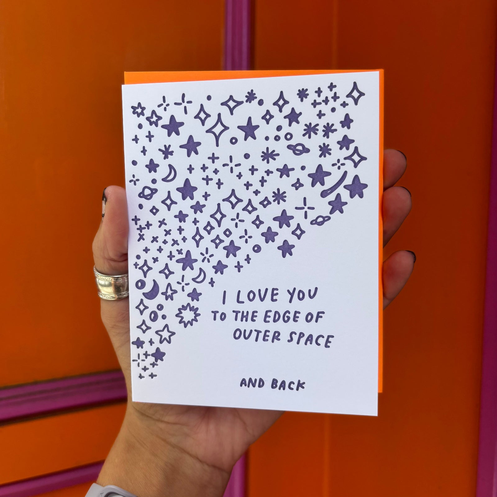 Someone holding the Outer Space I Love You Letterpress Greeting Card in front of an orange and pink wall.