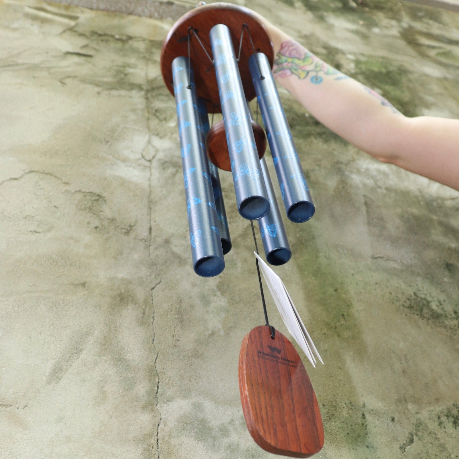 An up close image of someone holding the Butterfly Woodstock Garden Chime™ in front of a concrete wall.