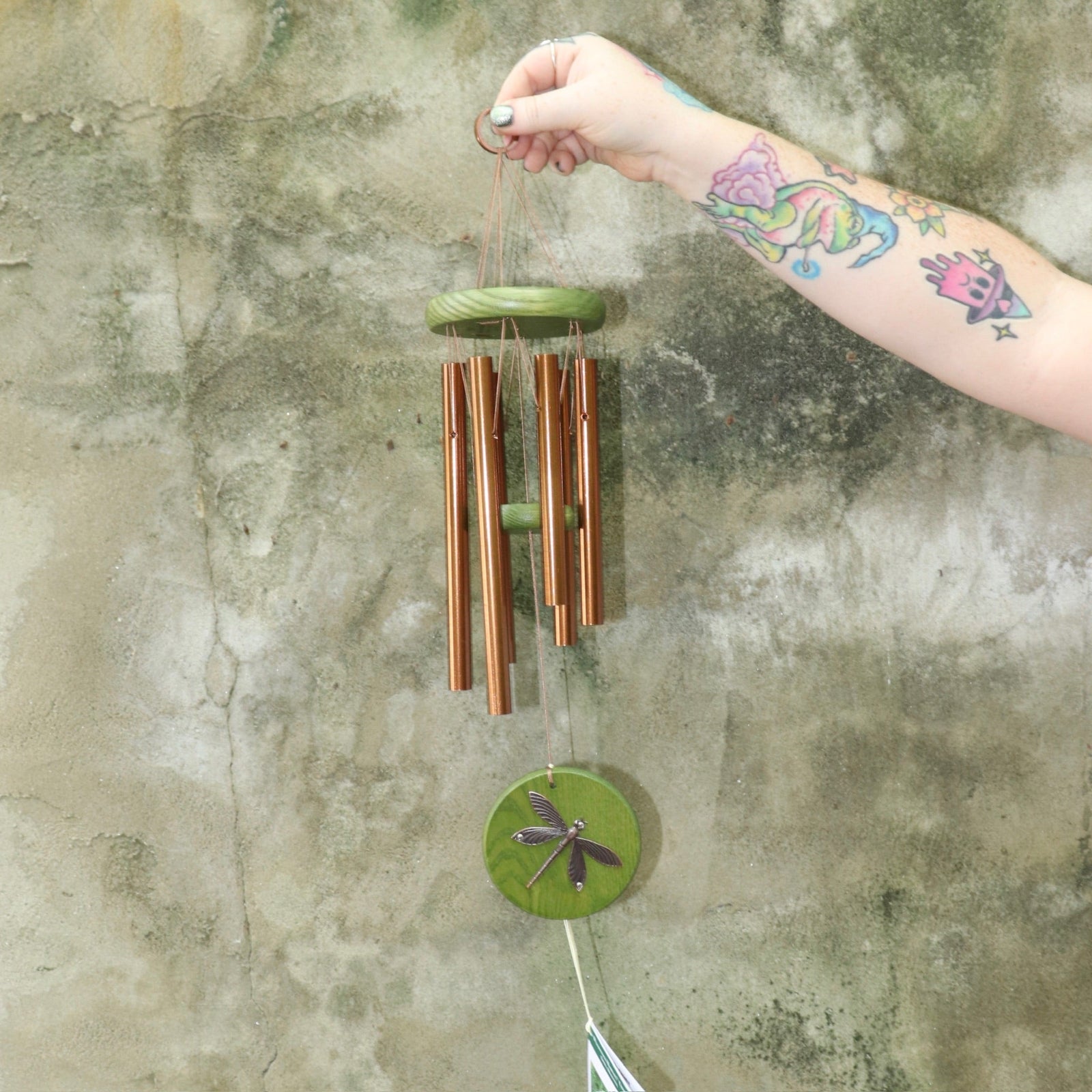 Someone holding the Green Dragonfly Woodstock Habitats™ Wind Chime in front of a concrete wall.