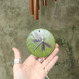 An up-close image of someone holding the Green Dragonfly Woodstock Habitats™ Wind Chime in front of a concrete wall.