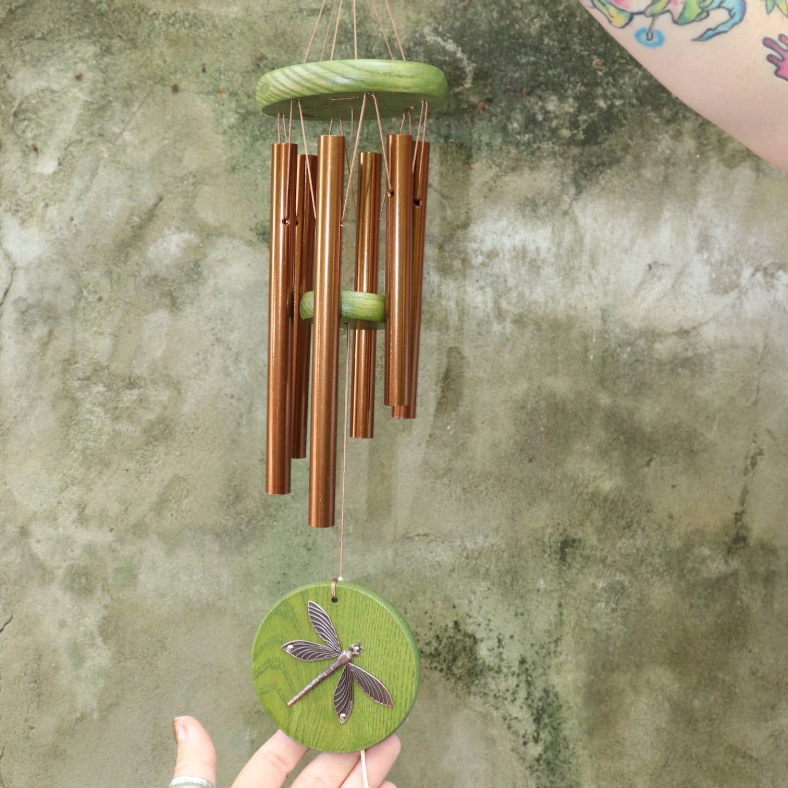 An up-close image of someone holding the Green Dragonfly Woodstock Habitats™ Wind Chime in front of a concrete wall.