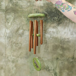 An up-close image of someone holding the Green Dragonfly Woodstock Habitats™ Wind Chime in front of a concrete wall.