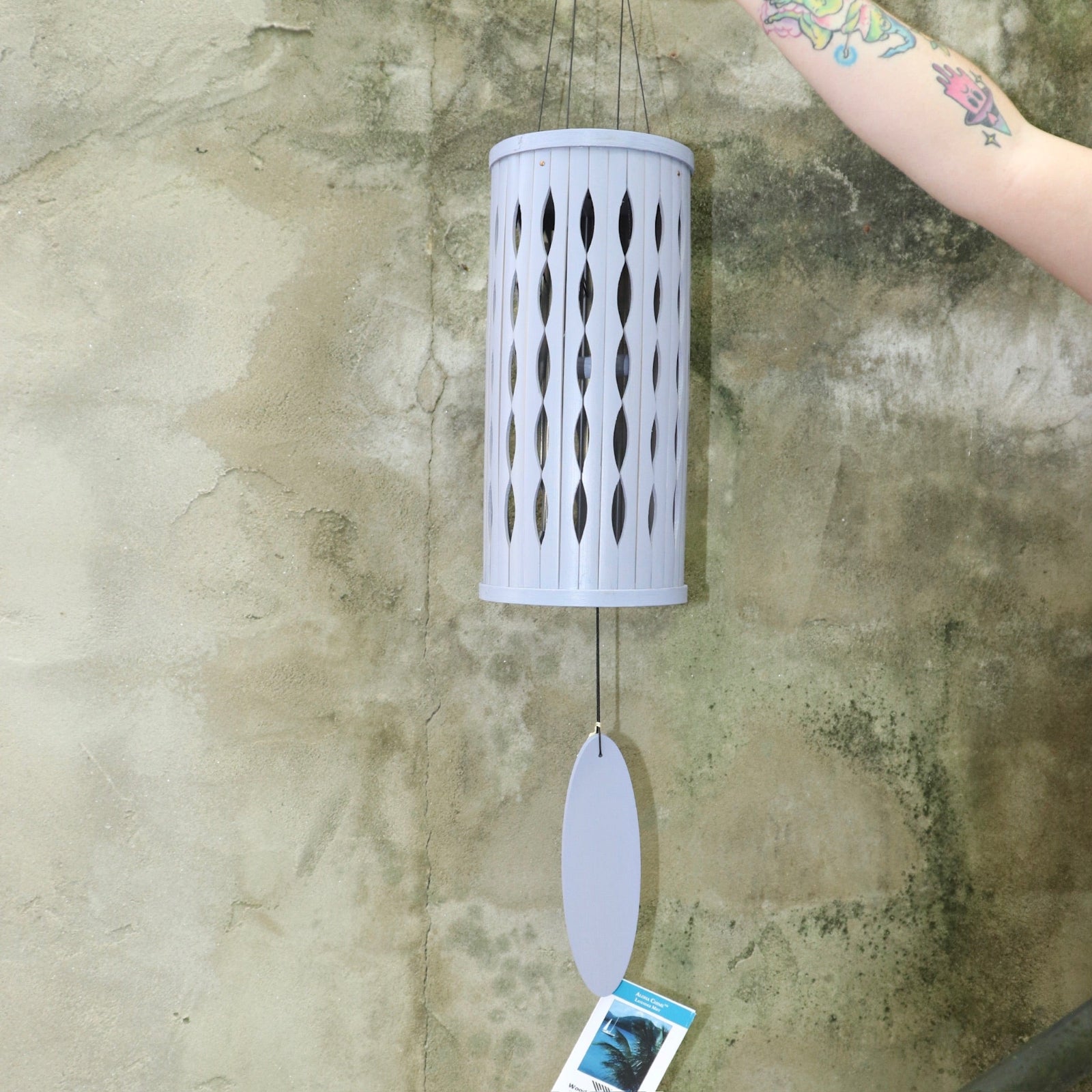 Someone holding the Lavender Mist Bamboo Aloha Wind Chime in front of a concrete wall.