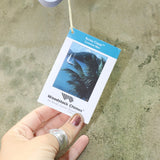 An up-close image of someone holding the tag of the Lavender Mist Bamboo Aloha Wind Chime in front of a concrete wall.