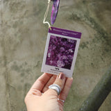 An up-close image of someone holding the tag of the Woodstock Amethyst Wind Chime in front of a concrete wall.