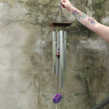 Someone holding the Woodstock Amethyst Wind Chime in front of a concrete wall.
