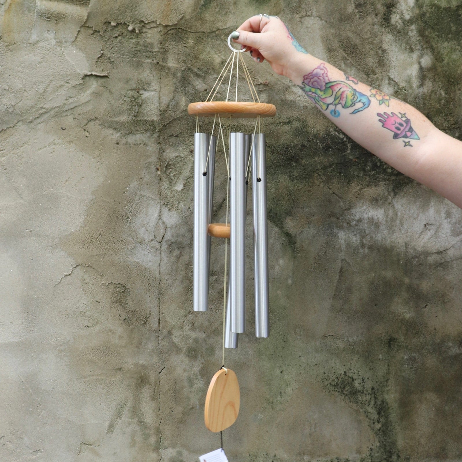 Someone holding the Woodstock Reflections Wind Chime in front of a concrete wall.