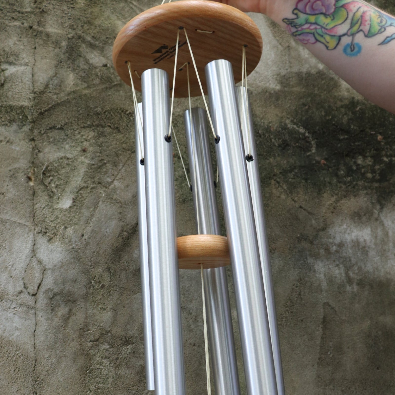 An up-close image of someone holding the Woodstock Reflections Wind Chime in front of a concrete wall.