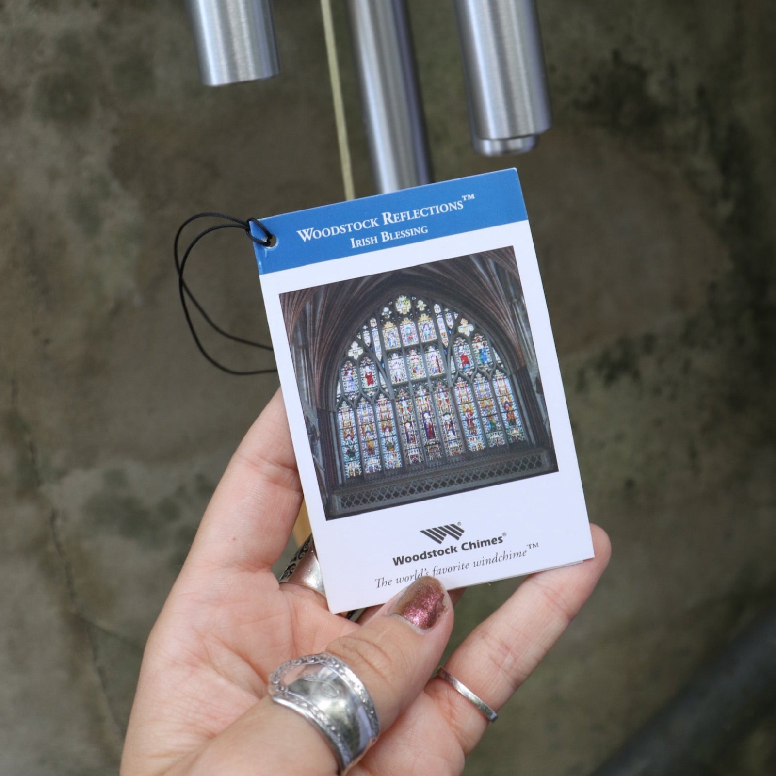 An up-close image of someone holding the tag of the Irish Blessing Woodstock Reflections Wind Chime in front of a concrete wall.