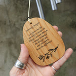 An up-close image of someone holding the Serenity Prayer Woodstock Reflections Wind Chime in front of a concrete wall.