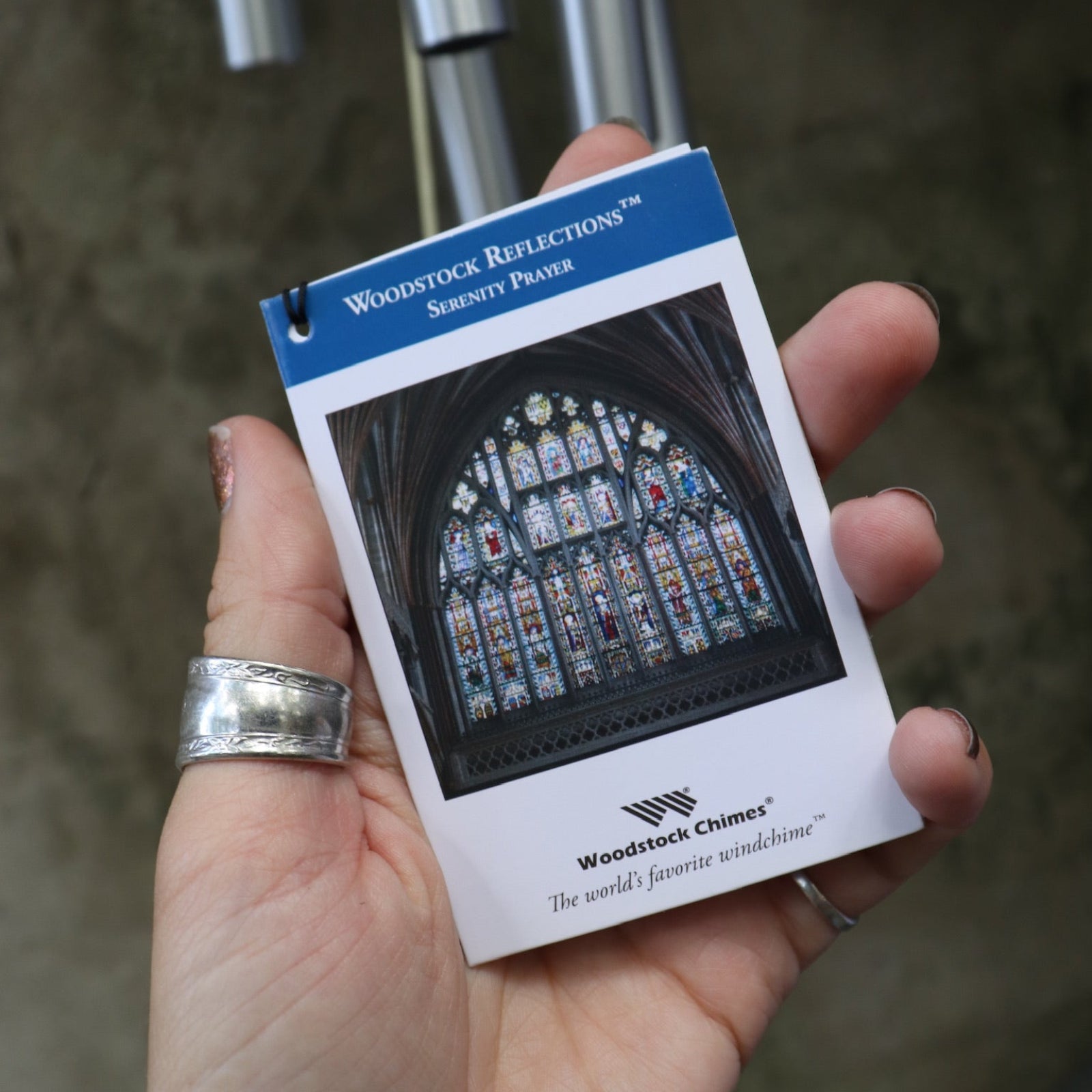 An up-close image of someone holding the Serenity Prayer Woodstock Reflections Wind Chime in front of a concrete wall.