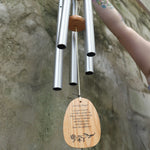 An up-close image of someone holding the Serenity Prayer Woodstock Reflections Wind Chime in front of a concrete wall.