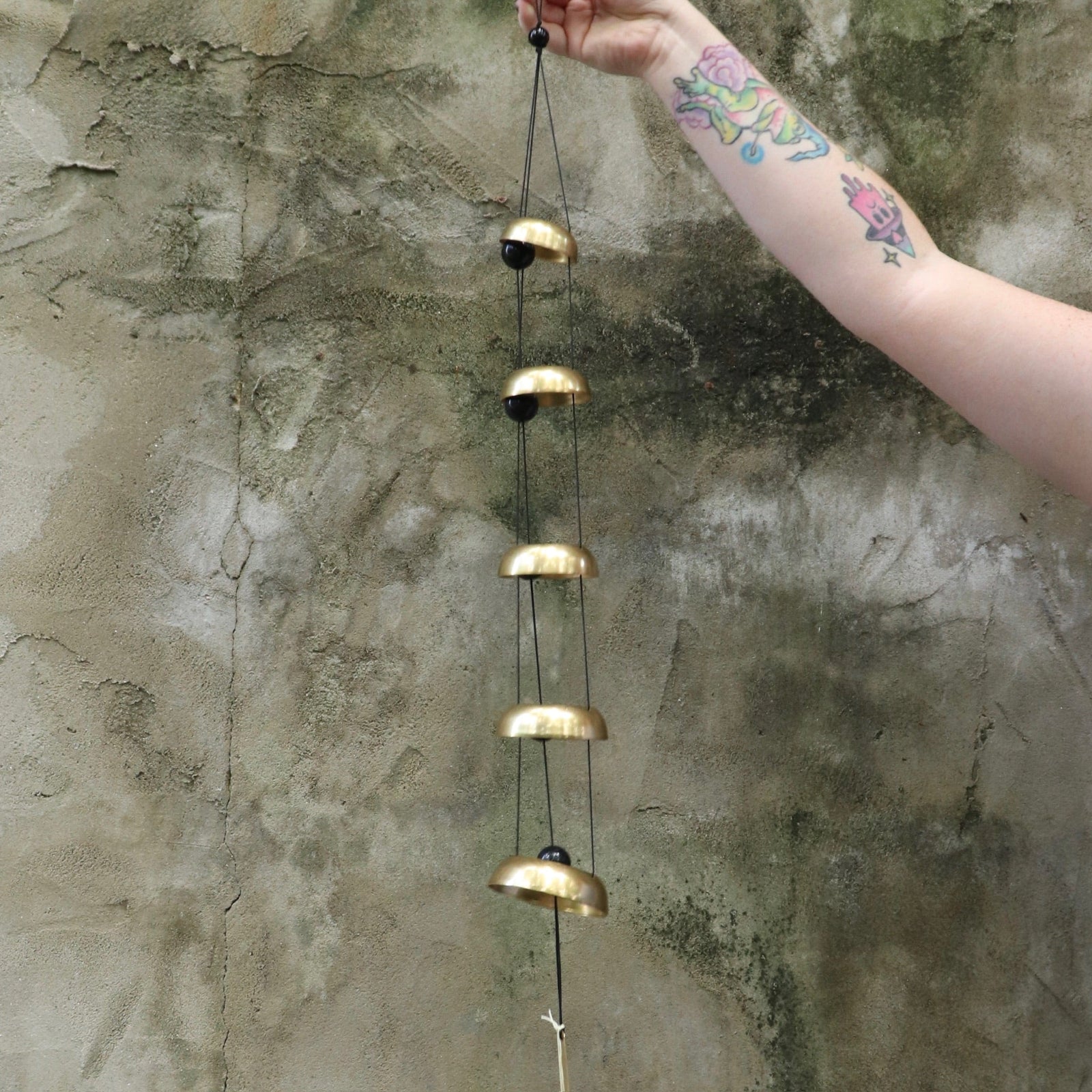 Someone holding the Brass Quintet Woodstock Temple Bells Wind Chime in front of a concrete wall.