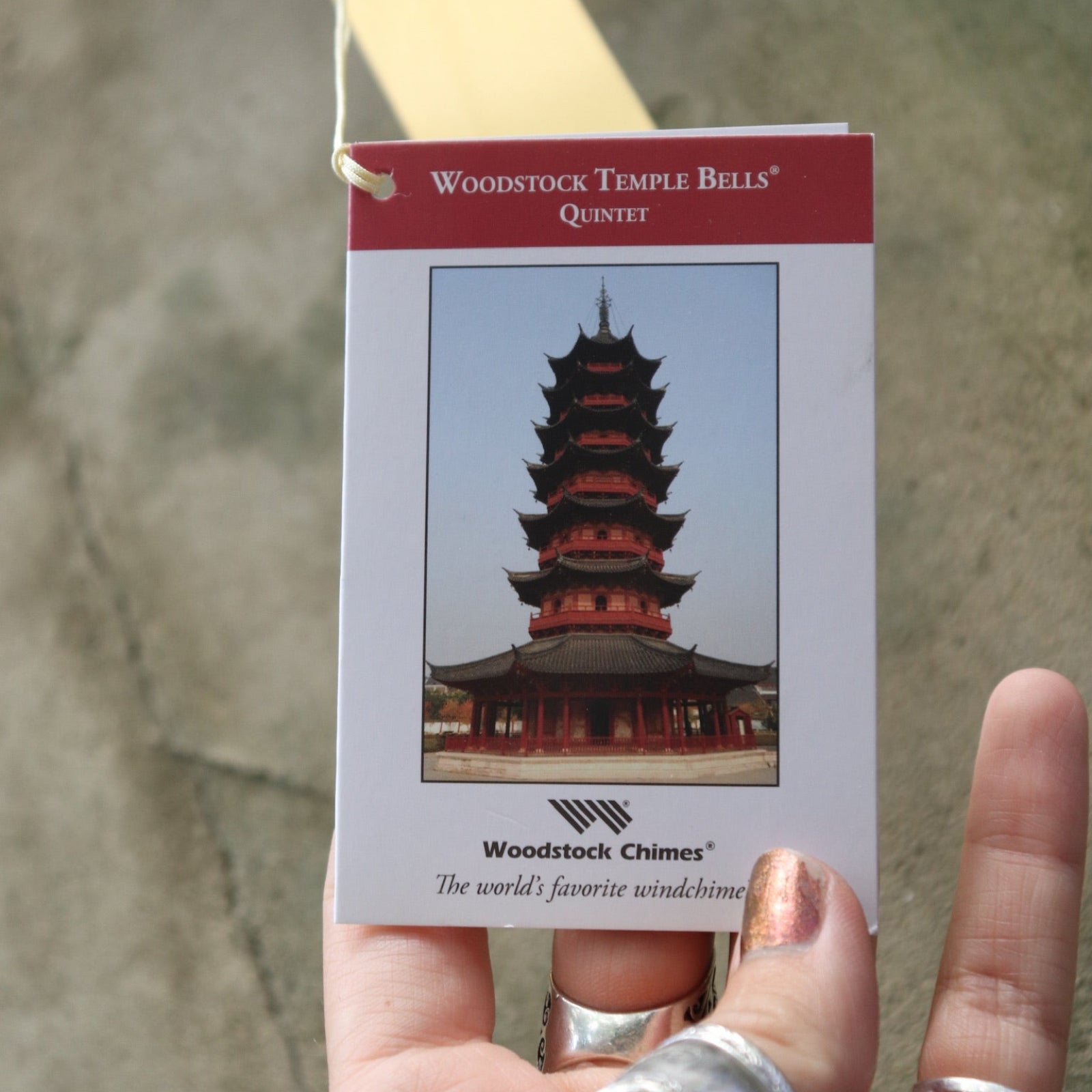 An up-close image of someone holding the tag of the Brass Quintet Woodstock Temple Bells Wind Chime in front of a concrete wall.
