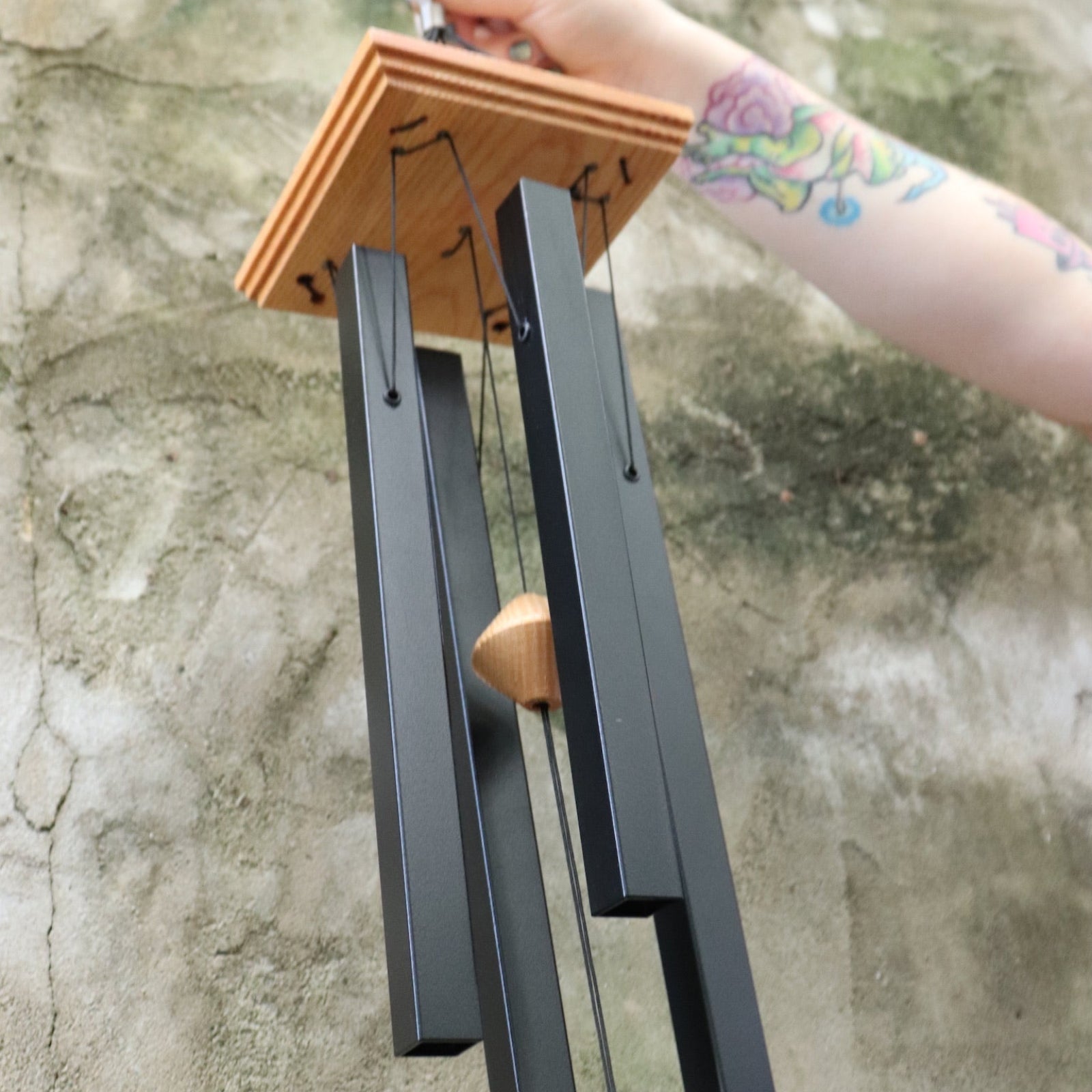 An up-close image of someone holding the Black Woodstock Craftsman Chime in front of a concrete wall.