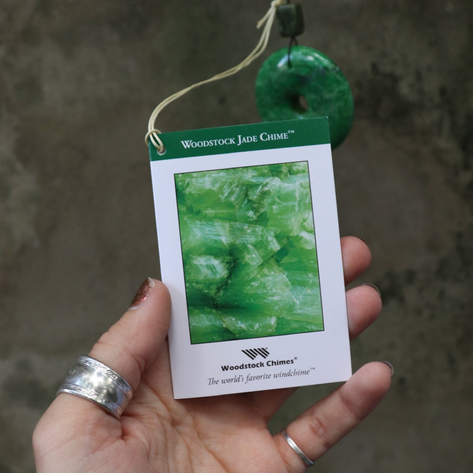 An up-close image of someone holding the tag of the Woodstock Jade Wind Chime in front of a concrete wall.
