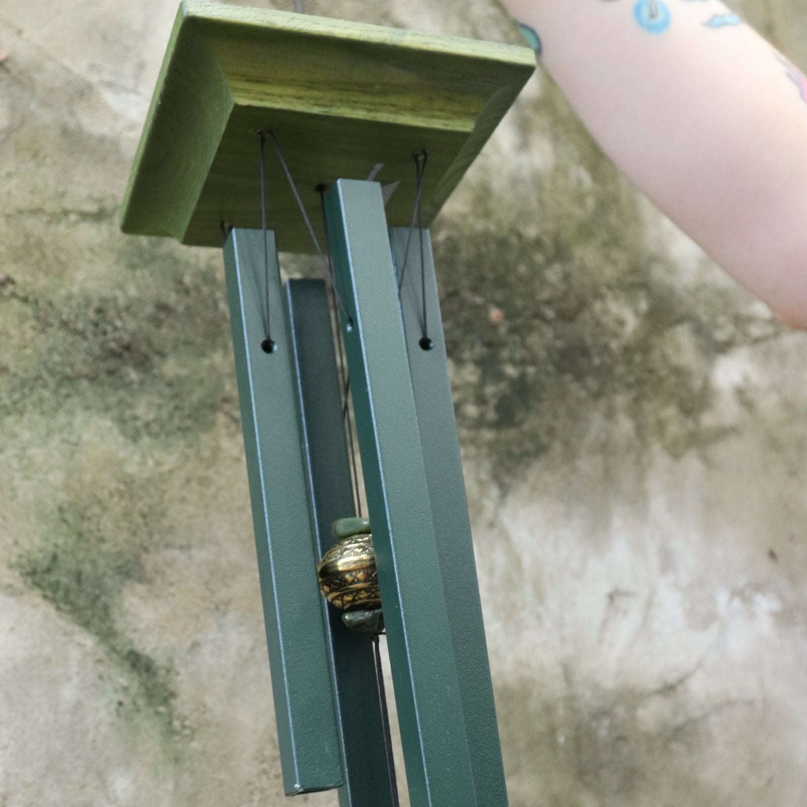 An up-close image of someone holding the Evergreen Woodstock Jade Wind Chime in front of a concrete wall.