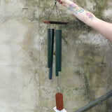 Someone holding the Evergreen Woodstock Craftsman Wind Chime in front of a concrete wall.