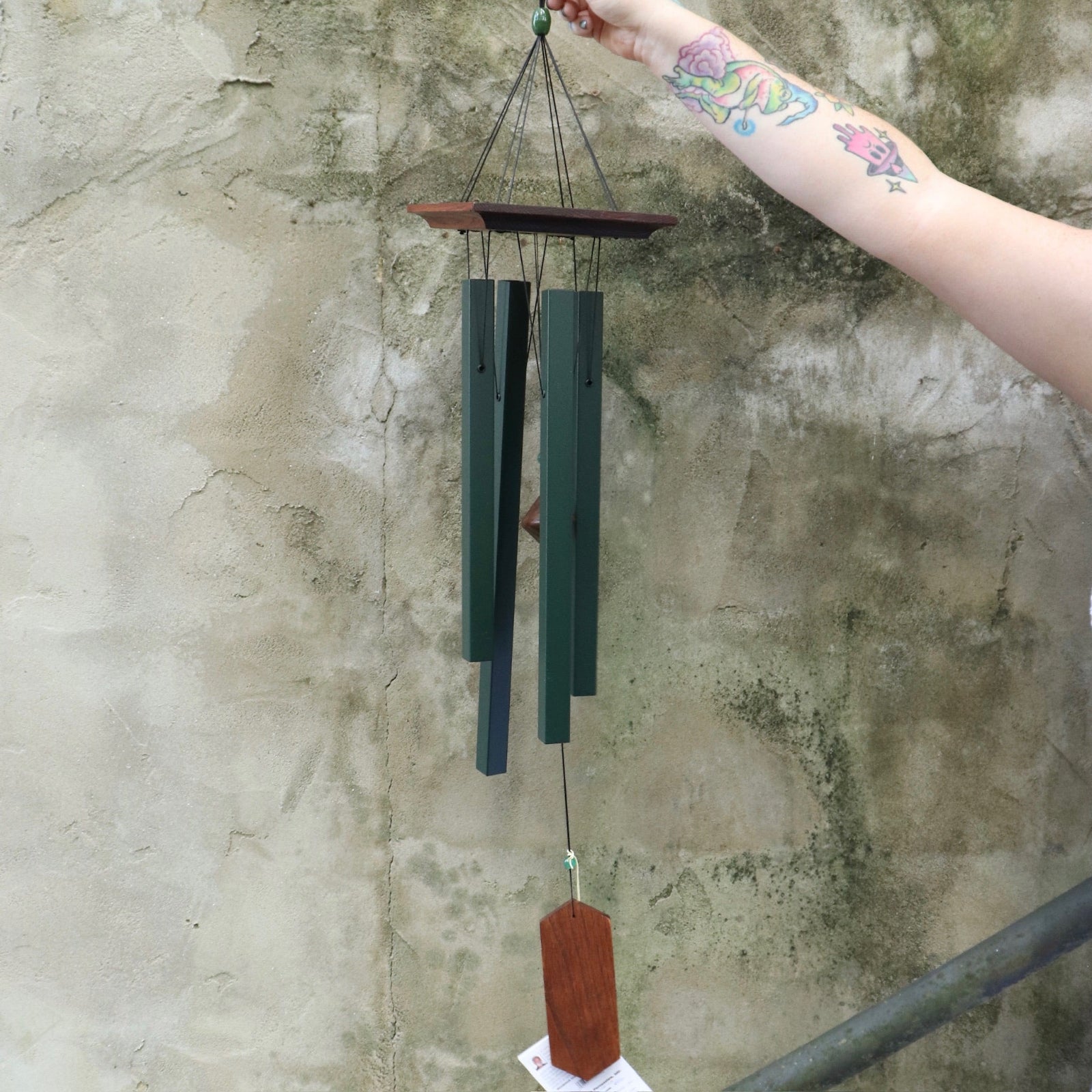 Someone holding the Evergreen Woodstock Craftsman Wind Chime in front of a concrete wall.