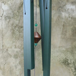 An up-close image of someone holding the Evergreen Woodstock Craftsman Wind Chime in front of a concrete wall.