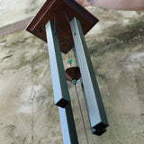 An up-close image of someone holding the Evergreen Woodstock Craftsman Wind Chime in front of a concrete wall.
