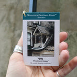 An up-close image of someone holding the tag of the Evergreen Woodstock Craftsman Wind Chime in front of a concrete wall.