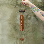Someone holding the Seven Stones Bronze Woodstock Chakra Chime in front of a concrete wall.