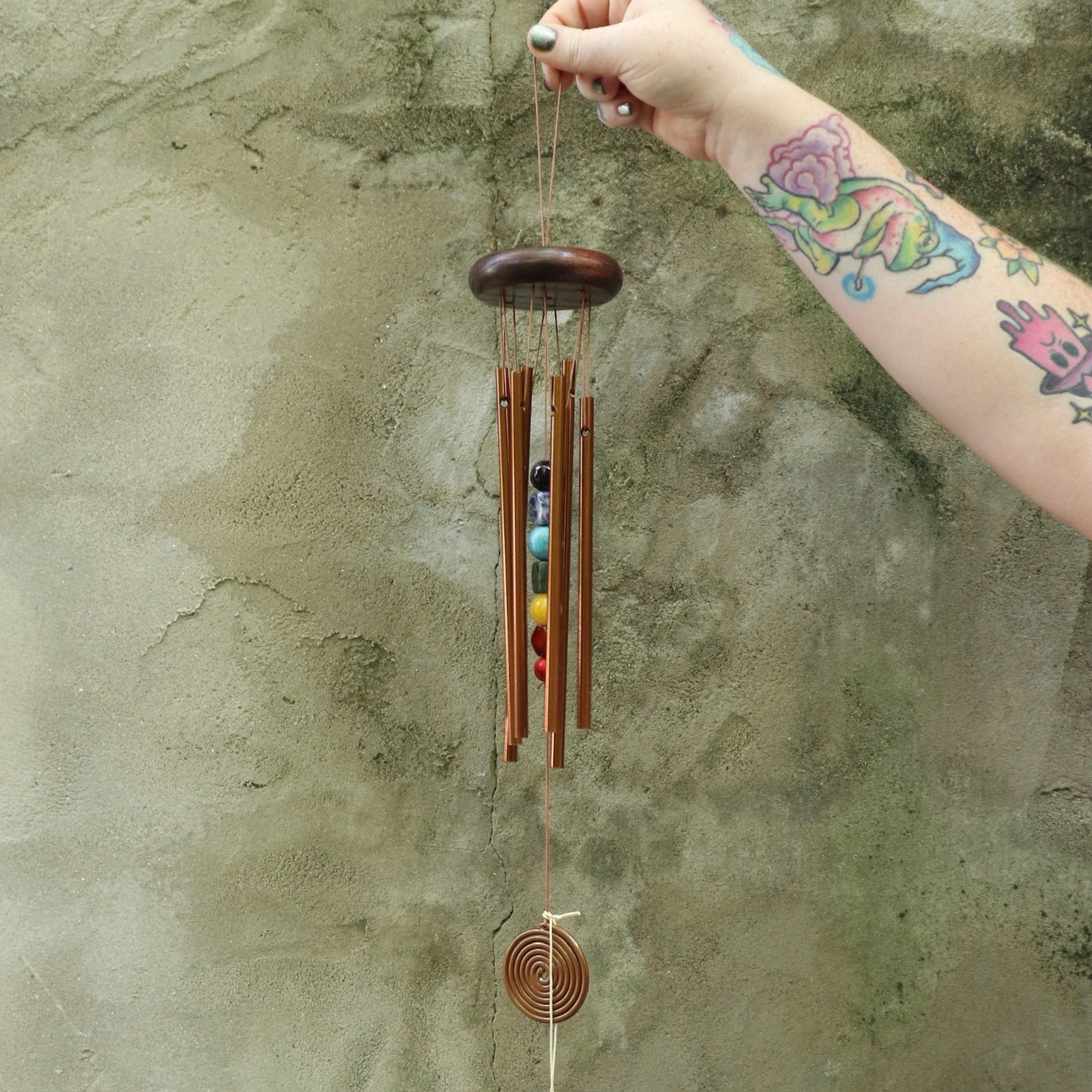 Someone holding the Seven Stones Bronze Woodstock Chakra Chime in front of a concrete wall.