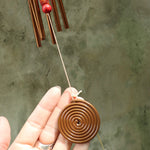 Someone holding the spiral clapper from the Seven Stones Bronze Woodstock Chakra Chime in front of a concrete wall.
